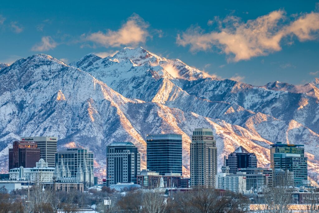 A city skyline with modern buildings set against the breathtaking backdrop of snow-capped mountains illuminated by the warm glow of a setting sun.