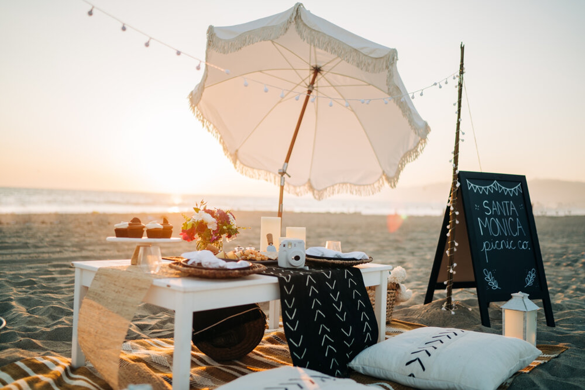 A serene beach picnic setup at sunset with an elegant white parasol, a low picnic table adorned with snacks, and a chalkboard sign indicating Los Angeles picnic co., all against the soft glow of a