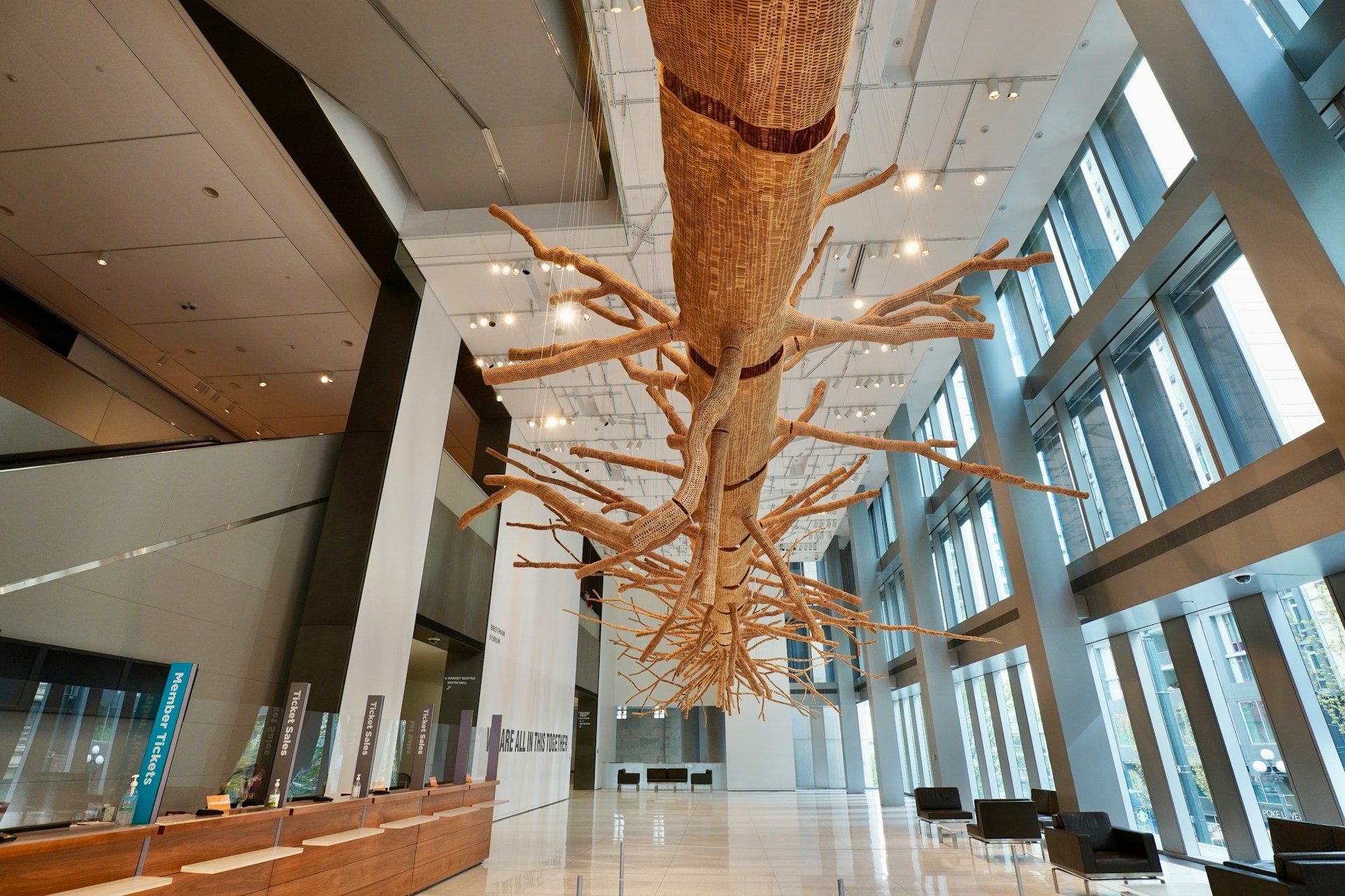 An upside-down tree sculpture hanging from the ceiling of a modern gallery space, creating a striking and thought-provoking centerpiece.
