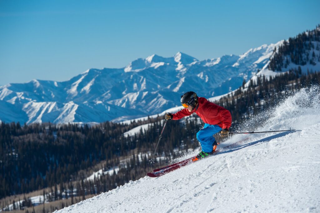 A skier in a red jacket carving through fresh snow with scenic mountain views in the background.