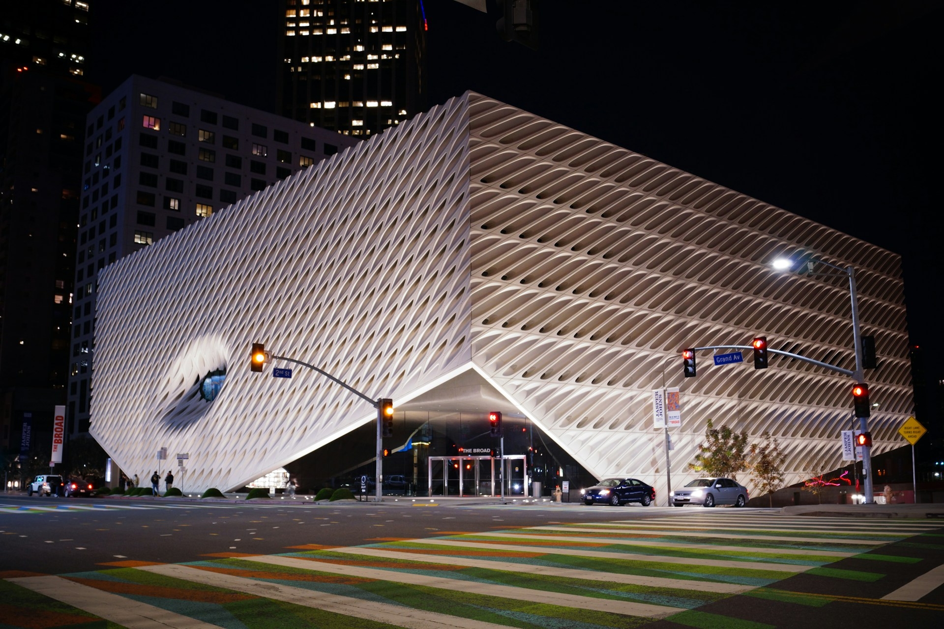 A nighttime view of a modern building in Los Angeles with a distinctive wavy facade, illuminated by city lights at a vibrant urban intersection.