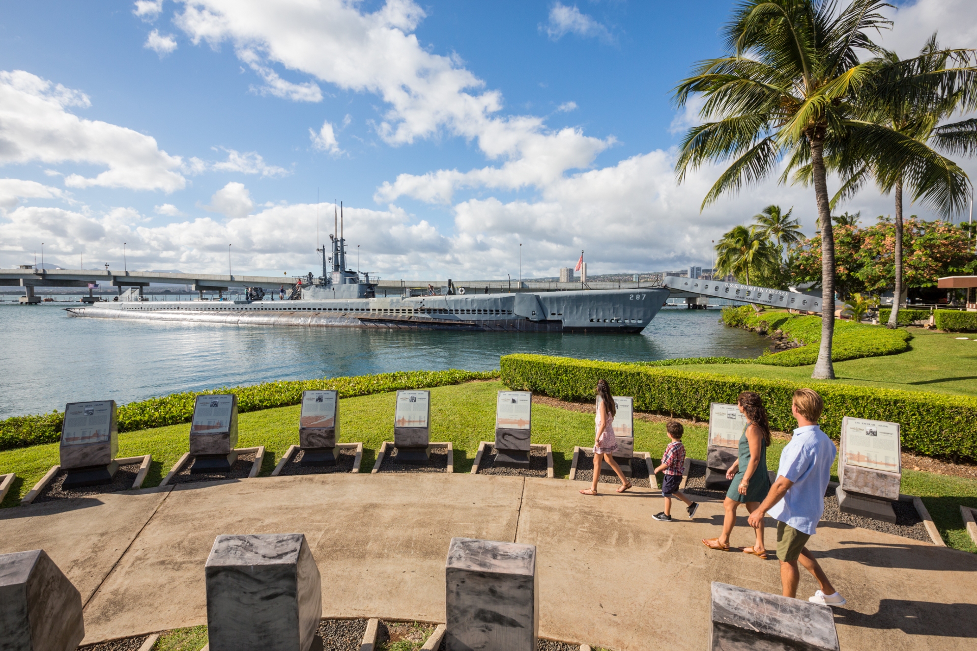 Visitors walking past informational plaques towards the uss bowfin submarine museum & park on a sunny day with clear skies and palm trees.