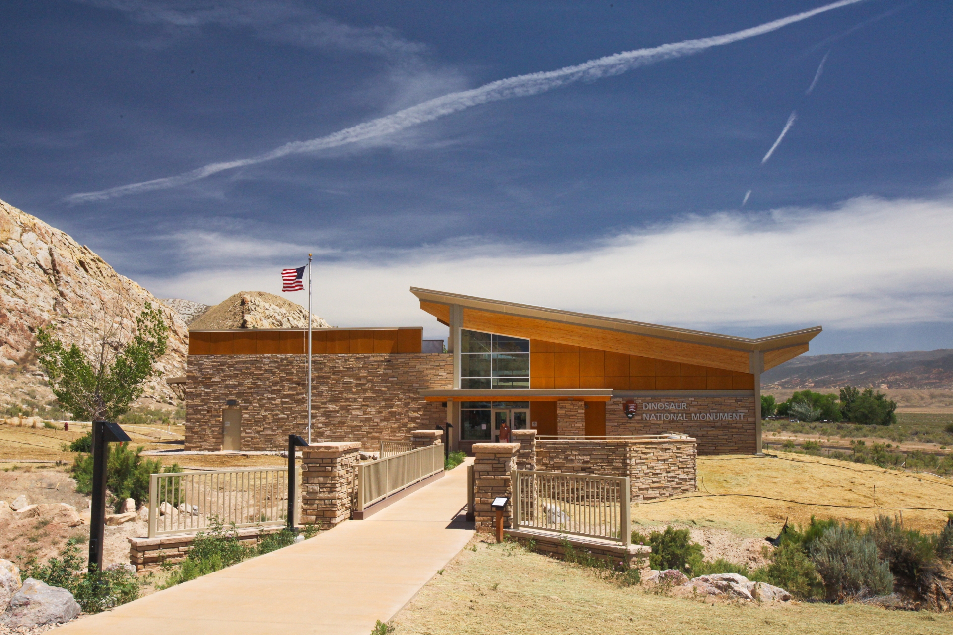 Modern visitor center nestled in a dry, hilly landscape under a blue sky with scattered clouds, featuring a flagpole with the american flag, stone and wood architectural elements, and a paved access path.