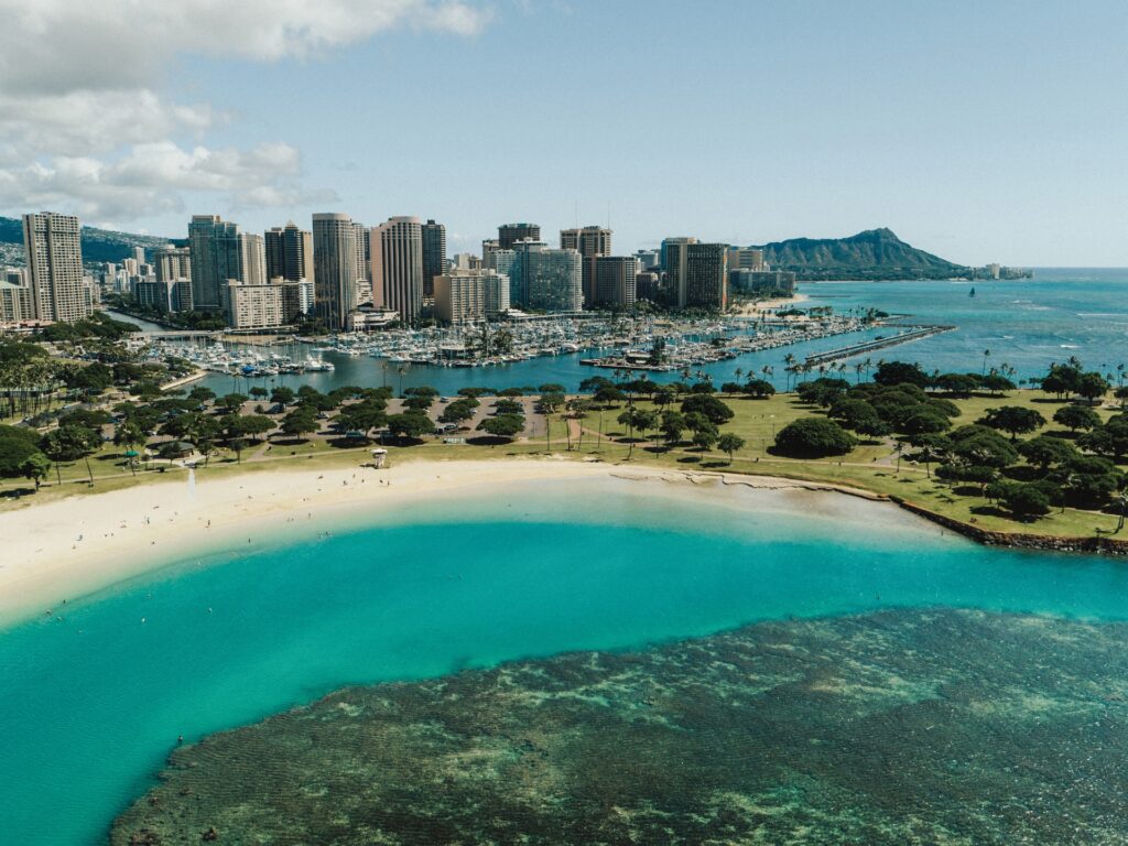 An aerial view of a pristine beachfront with crystalline waters, surrounded by a lush park, with a bustling city skyline and a majestic mountain in the distance.
