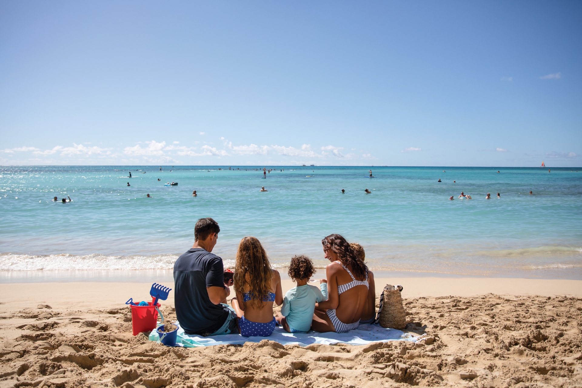 A family sitting together on the sand, enjoying a sunny day at the beach while watching swimmers in the crystal-clear sea.