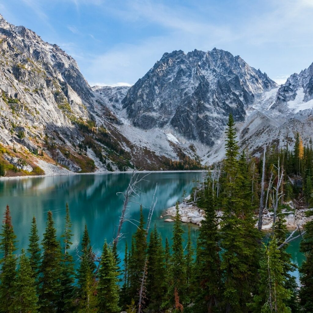 Majestic mountains rise above a tranquil turquoise lake, surrounded by dense evergreen trees.