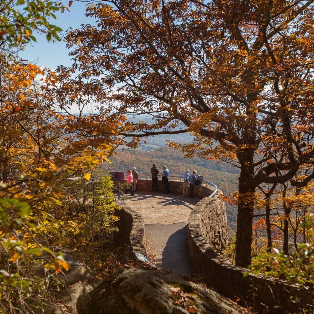 Visitors on a stone overlook surrounded by autumn foliage, with distant hills visible.